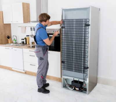 Serviceman In Overall Working On Fridge With Screwdriver In Kitchen