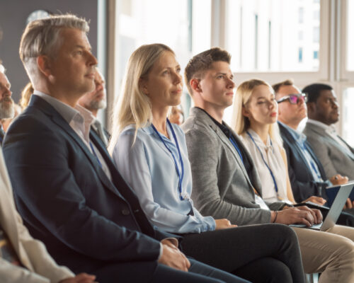 Caucasian Woman Sitting in Crowded Audience at Human Resource Conference. Female Delegate Focused On Presenter. HR Manager Listening to Inspirational Entrepreneurship Presentation About Social Studies
